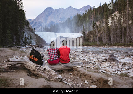 Couple sitting on log near waterfall Stock Photo