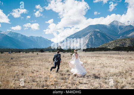 Bride and groom running away with backs to camera in a field with a mountain backdrop on a perfect summer day with blue sky and big white clouds Stock Photo