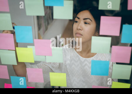 Female executive sticking adhesive notes on glass wall Stock Photo