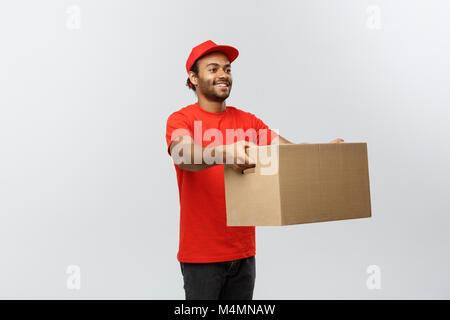 Delivery Concept - Portrait of Happy African American delivery man in red cloth holding a box package. Isolated on Grey studio Background. Copy Space. Stock Photo