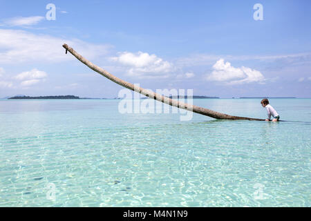 Young caucasian blonde boy in bathing suit climbing on long palm tree trunk over sandy beach and turquoise water Stock Photo