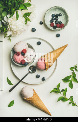 Flat-lay of Pink strawberry and coconut ice cream scoops in bowl, sweet waffle cones, peony flowers bouquet and fresh berries over white background, t Stock Photo