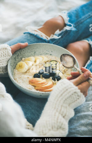 Healthy winter breakfast in bed. Woman in woolen sweater and shabby jeans eating vegan almond milk oatmeal porridge with berries, fruit and almonds, c Stock Photo