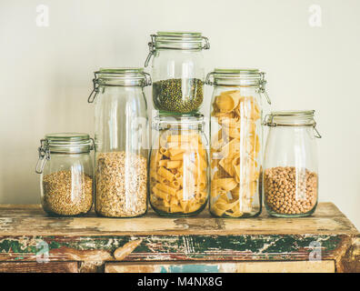 Various uncooked cereals, grains, beans and pasta for healthy cooking in glass jars on rustic wooden table, white background. Clean eating, vegetarian Stock Photo