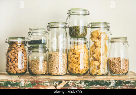 Various uncooked cereals, grains, beans and pasta for healthy cooking in glass jars on wooden table, white background, horizontal composition. Clean e Stock Photo