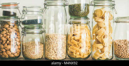 Various uncooked cereals, grains, beans and pasta for healthy cooking in glass jars on wooden table, white background, close-up. Clean eating, vegan, Stock Photo