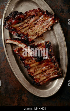 Closeup shot of flavorful grilled pork ribs in thick barbeque sauce cut in messy way served in vintage metal tray on dark textured background. Pub sty Stock Photo