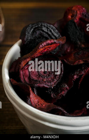 Organic homemade beetroot chips cooked in oven with olive oil, pepper and salt in white ceramic bowl on wooden background. Vertical composition, close Stock Photo