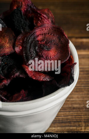 Organic homemade beetroot chips cooked in oven with olive oil, pepper and salt in white ceramic bowl on wooden background. Vertical composition, close Stock Photo
