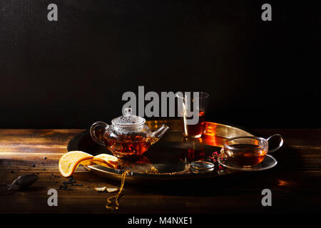 Small glass teapot and glasses with hot black tea, dried rose petals, pocket magnifier on golden chain, squeezed orange slice on golden tray. Evening Stock Photo