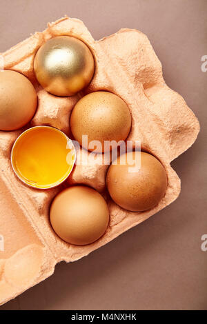 Horizontal closeup shot with colorful cardboard container of pink color with chicken eggs, one yolk visible, one egg painted golden. Easter concept Stock Photo