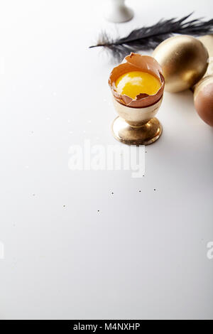Easter holiday composition with eggs painted golden, yolk in egg stand, black feather and golden ribbon. White background, copy space Stock Photo