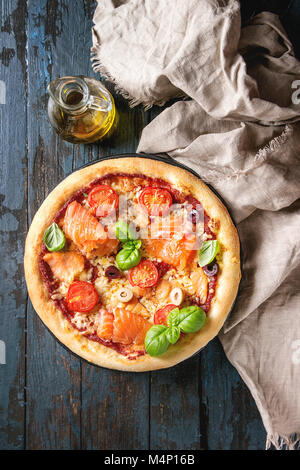 Traditional pizza with smoked salmon, cheese, tomatoes and basil served on black plate with textile and olive oil over dark blue wooden background. To Stock Photo