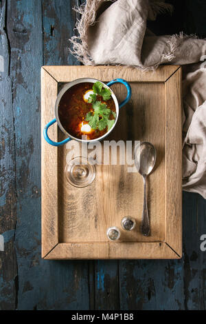 Pan of traditional beetroot borscht soup with sour cream and fresh coriander served with glass of vodka on wood tray over dark blue wooden background. Stock Photo