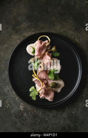 Vitello tonnato italian dish. Thin sliced veal with tuna sauce, capers and coriander served on black plate over old dark metal background. Top view, s Stock Photo