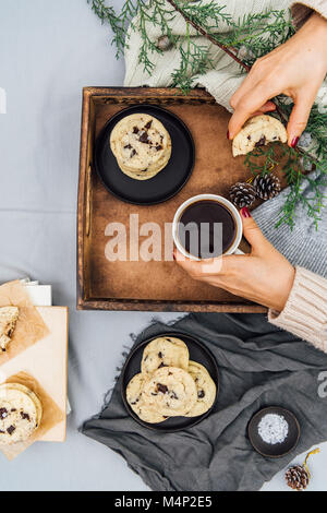 A woman holding a cup of coffee and a half eaten cookie in a breakfast tray  photographed from top view. More cookies on a black plate and on some boo Stock Photo