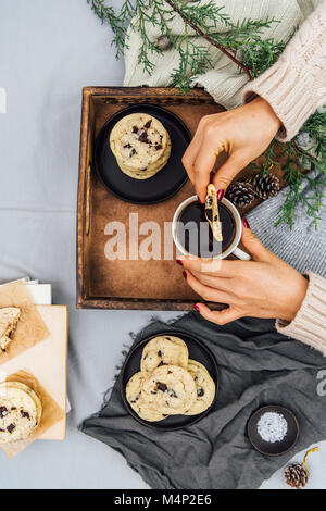 A woman dipping a chocolate chip cookie into her coffee in a wooden tray photographed from top view. More cookies on a black plate and on some books, Stock Photo