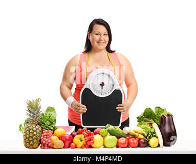 Overweight woman with a weight scale and a measuring tape behind a table with vegetables and fruit isolated on white background Stock Photo