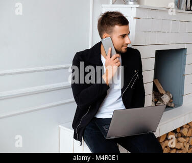 Casual bearded man in black clothes using laptop near fireplace. Stock Photo