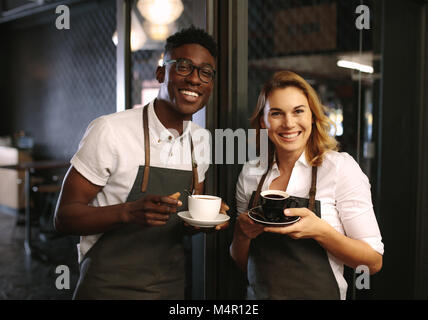 Male and female barista standing at the entrance of coffee shop holding coffee cups. Happy coffee shop owners posing with coffee cups in hand. Stock Photo