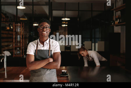 Man standing in his coffee shop with arms crossed while woman is working in the background. Young entrepreneurs managing their coffee shop.
