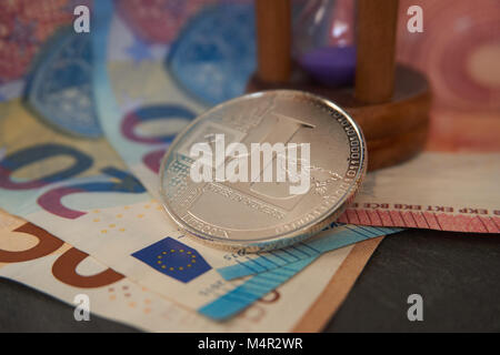 Silver litecoin and hourglass  lying on the  euro notes, grey stone background. Cryptocurrency digging speed concept, selective focus. Stock Photo