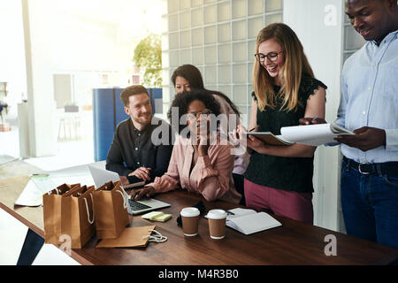 Diverse group of smiling colleagues talking together while busy working at a table in a modern office Stock Photo