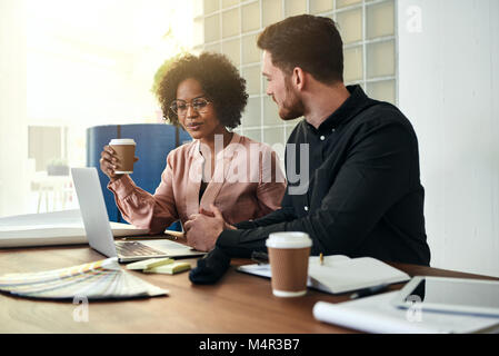 Two designers sitting at a table in a modern office talking together and working with a laptop Stock Photo