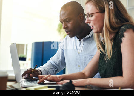 Two focused business colleagues sitting at a table in an office talking together and working online with a laptop Stock Photo