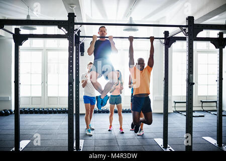 Two fit young men having a friendly pullups competition in a gym with a diverse group of friends cheering in the background Stock Photo
