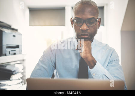 Focused African businessman deep in thought while sitting at a desk working online with a laptop from his home office Stock Photo