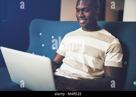 Young African male entrepreneur smiling while sitting on a sofa working online with a laptop Stock Photo