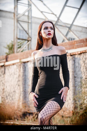 Portrait of the beautiful goth girl (informal model) in black dress and tights standing in industrial area Stock Photo