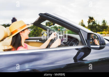 Close side view of young lady driver in an open modern sports convertible car with soft top folded down against clear sky. Stock Photo