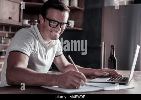 Joyful millennial guy taking notes while working on laptop Stock Photo
