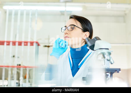 Waist-up portrait of pensive young scientist wearing eyeglasses and white coat looking away while preparing report about conducted experiment, interio Stock Photo
