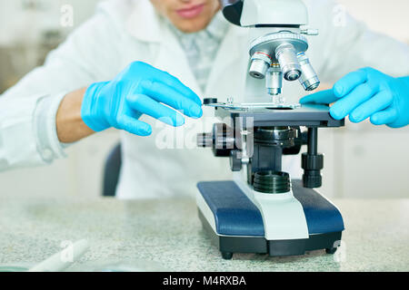 Close-up shot of unrecognizable male researcher wearing white coat and rubber gloves examining sample with microscope while sitting at laboratory desk Stock Photo