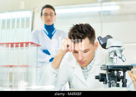 Concentrated young researcher wearing white coat leaning head on hand while sitting at desk, his pretty female colleague standing behind him, interior Stock Photo