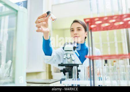 Confident young researcher wearing white coat sitting at desk and inspecting sample of in vitro meat, microscope and laboratory glassware on foregroun Stock Photo