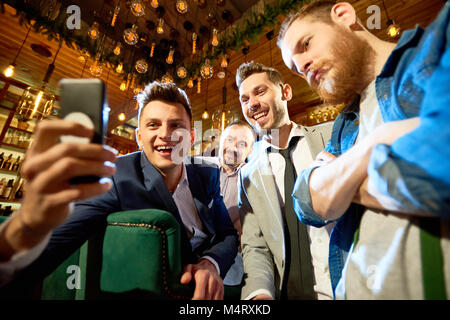 Smiling bearded friends taking selfie with help of smartphone while having long-awaited gathering in modern pub Stock Photo