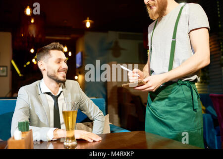 Cheerful bearded man wearing stylish suit sitting at cafe table and making order, red-haired waiter wearing apron standing next to him and taking note Stock Photo