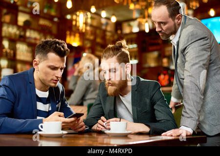 Handsome young manager using smartphone while having informal working meeting with colleagues, interior of modern cafe on background Stock Photo