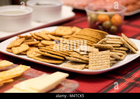 Crackers on a white tray, cheeses on a clear glass tray, dip in two white bowls, all on a red tartan tablecloth Stock Photo