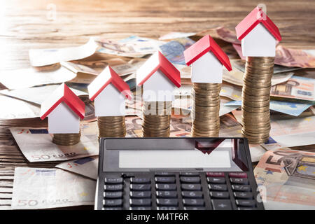 Miniature Houses Resting On Increasing Coin Stack With Calculator On Euro Notes Stock Photo
