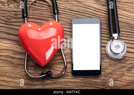 Red Heart Over The Stethoscope With Blank Screen Smart Phone On Wooden Table Stock Photo