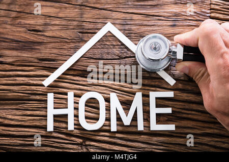 Close-up Of A Person Hand Holding Stethoscope Near Home Text On Wooden Table Stock Photo
