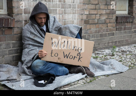 Homeless Man In Hood Sitting On Street Holding Cardboard With Text Willing To Work Stock Photo