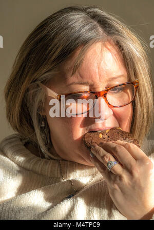 Closeup of an attractive, caucasian, short-haired, middled aged woman, wearing glasses, taking a bite from a home baked cookie. England, UK. Stock Photo