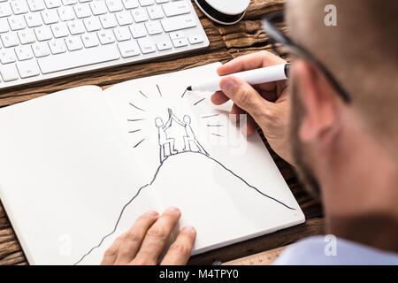 Businessman Drawing The Figures Giving High Five On Top Of Mountain In Notebook Stock Photo