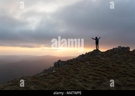 Silhouette of young woman at sunset raising hands, standing on the top of the mountain with a view over the barren grass covered mountain landscape Stock Photo